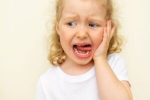 Little girl in white shirt holding her hand to her face and sticking her tongue out in pain